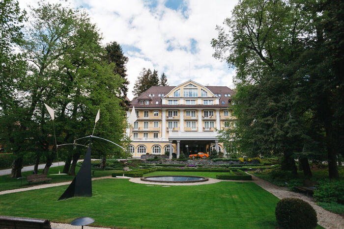 A grand hotel with celebrity relatives on a lush green lawn featuring a modern sculpture and a circular fountain under a cloudy sky.