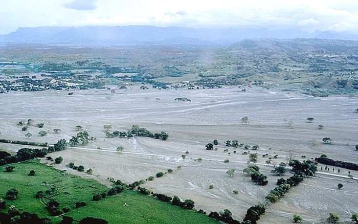 Disturbing d***h stories: Aerial view of a landscape devastated by a volcanic eruption, grey ash covering fields and a riverbed.