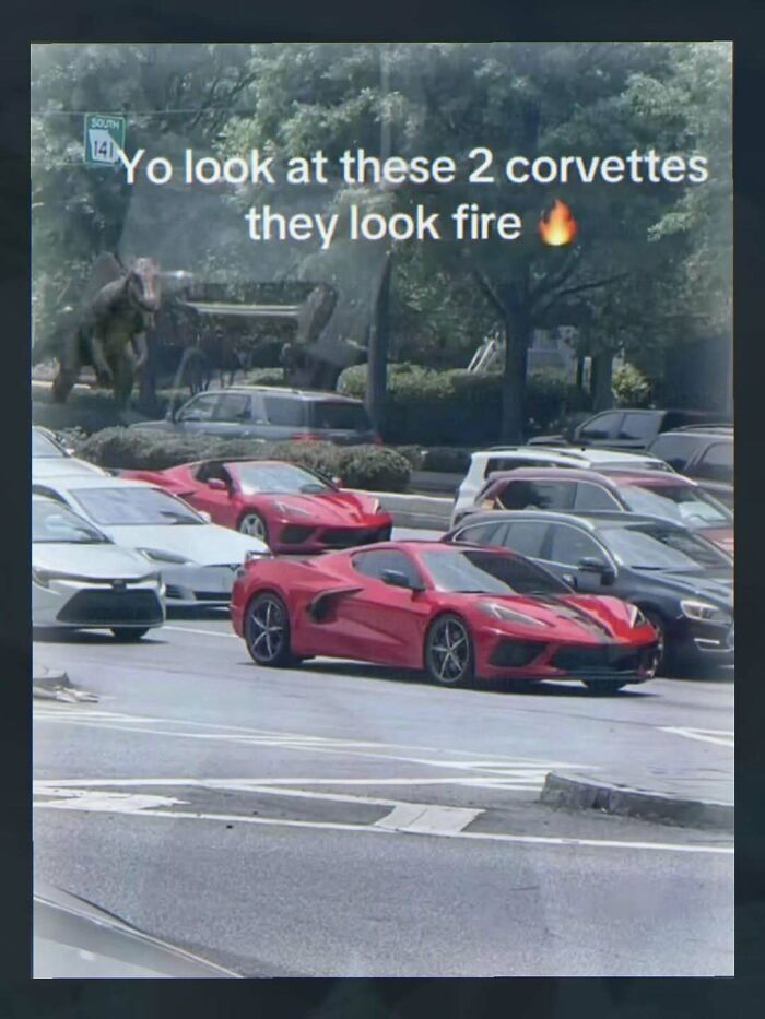 Humorous photo of two red Corvettes in parking lot called fire with dinosaur in background