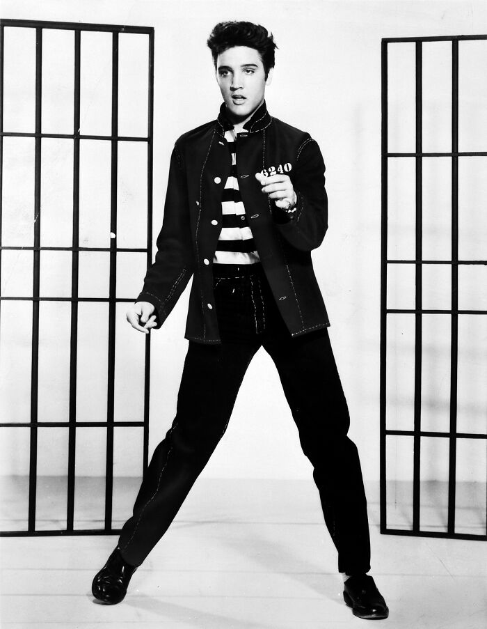 Black and white photo of a man in prison uniform holding a number card in front of jail bars