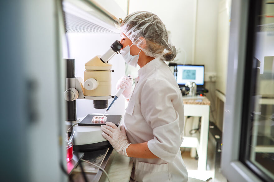 Scientist in a lab coat and mask using a microscope and pipette for a scientific breakthrough experiment.