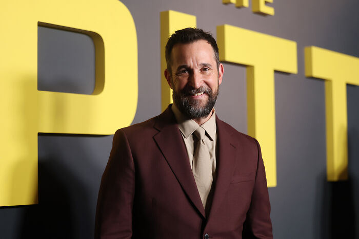Noah Wyle sorrindo em terno marrom no evento promocional do show The Pitt