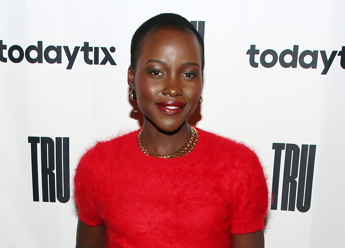 Lupita Nyong'o smiling in red dress at a media event related to The Odyssey casting remark