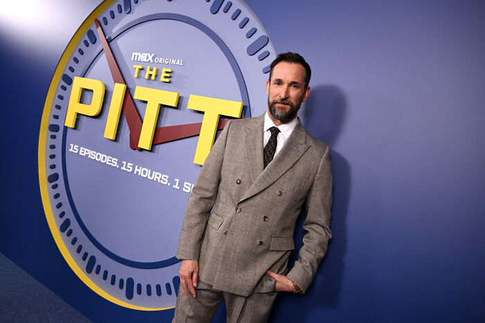 O ator Noah Wyle posando no evento de estreia da série The Pitt em terno cinza