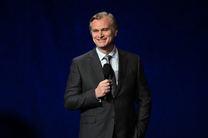 Christopher Nolan sorrindo, segurando um microfone, contra um fundo azul escuro. Os fãs reagiram brutalmente ao trailer do Odyssey.
