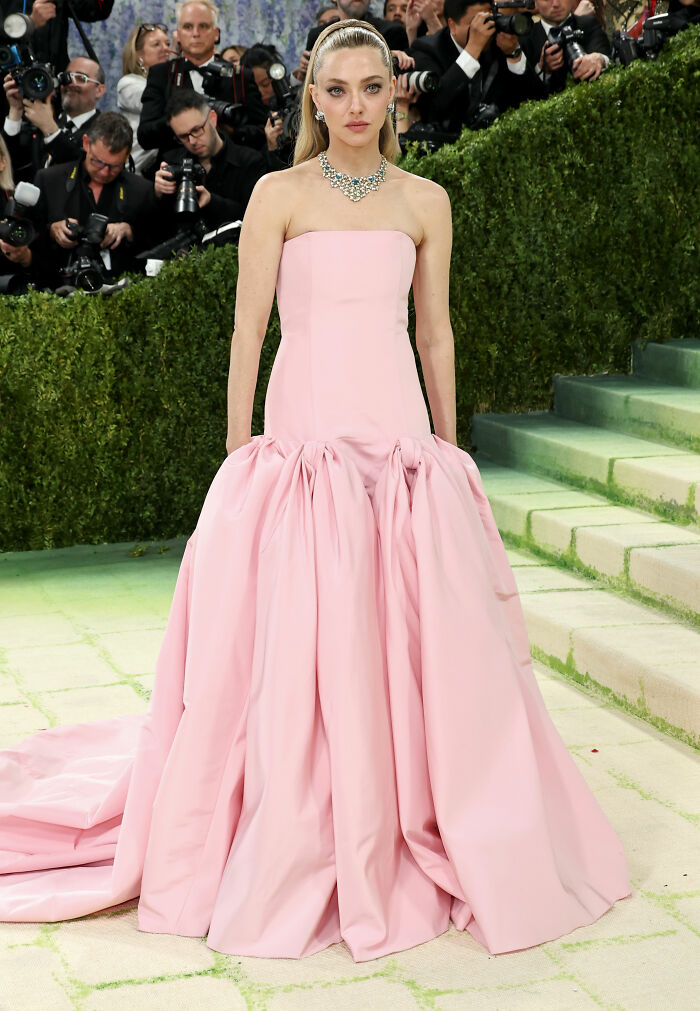 A celebrity poses in a stunning pink gown with a ruffled skirt and ornate necklace, paparazzi visible in background. MET Gala outfits.