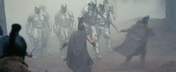 Personagens em trajes gregos antigos confrontam figuras blindadas em uma floresta enevoada, evocando A Odisseia de Christopher Nolan.