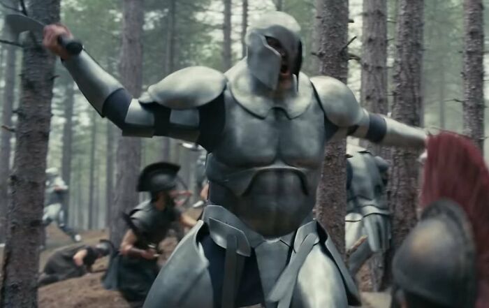Um guerreiro fortemente blindado, com um capacete cobrindo o rosto, empunha uma espada em uma batalha na floresta pela Odisséia.