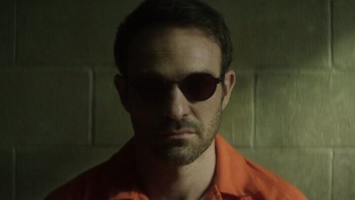 Charlie C*x como Demolidor, usando óculos e terno laranja, em um cenário mal iluminado para Born Again.
