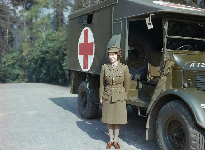 Woman in military uniform standing next to vintage ambulance with red cross symbol