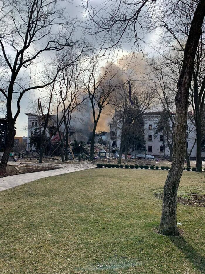 A truly disturbing d***h story scene: a bombed-out building spews smoke into a grey sky, framed by bare trees and a park.