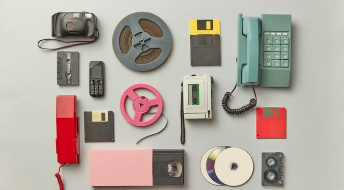 A nostalgic flat lay of childhood picks: a camera, cassette tapes, floppy disks, cell phone, VCR, and old telephones.