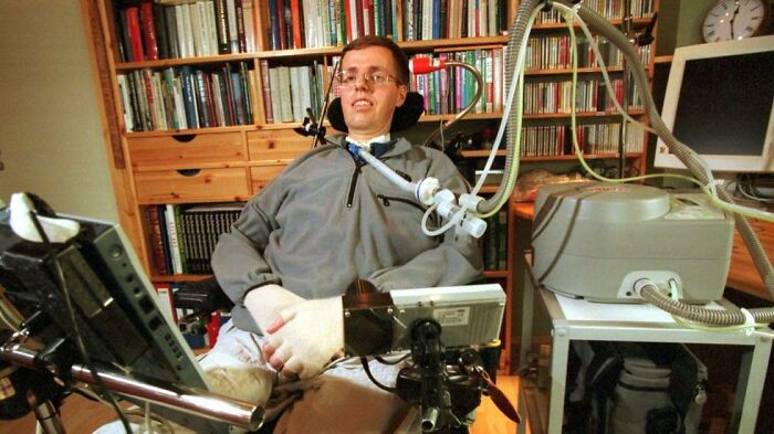 A man with a tracheostomy tube and glasses sits in a wheelchair, surrounded by medical equipment. Disturbing d***h stories.