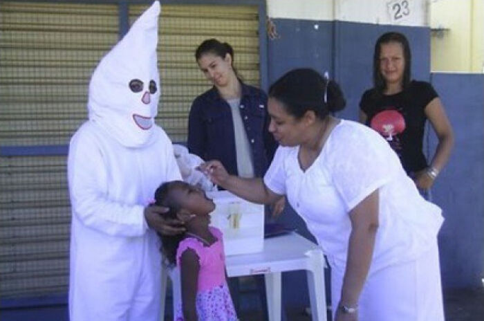 Woman in white administering medicine to child while person in white costume holds the child