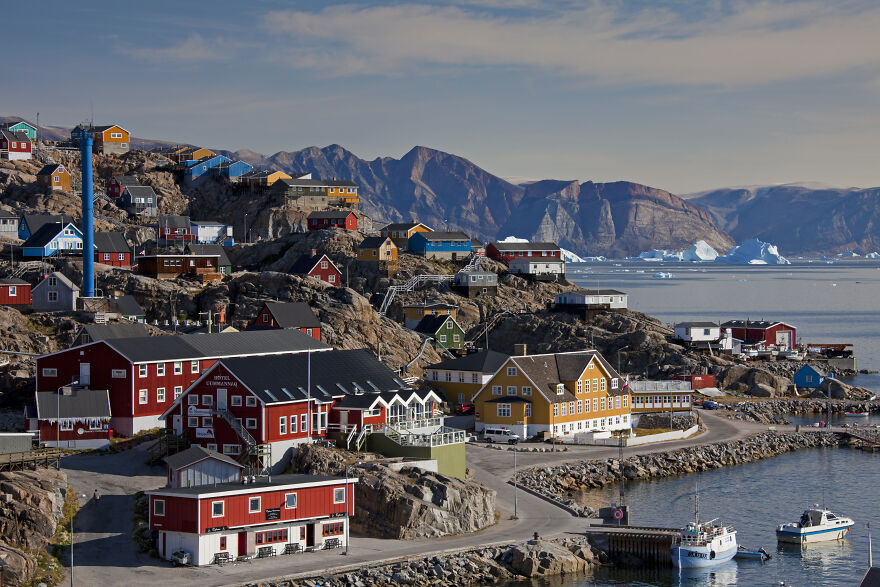 Colorful houses line a rocky coast in one of Earth's most isolated places, with mountains and icebergs in the distance.