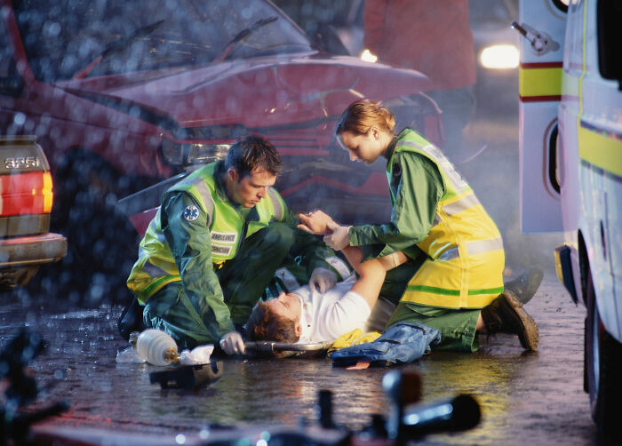 Paramedics aiding an injured patient on a stretcher at a car accident scene, highlighting doctors asking about being alive.