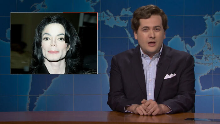 An SNL host at a news desk, with a picture of Michael Jackson beside him, mocking Met Gala outfits. An SNL host at a news desk, with a picture of Michael Jackson beside him, mocking Met Gala outfits.