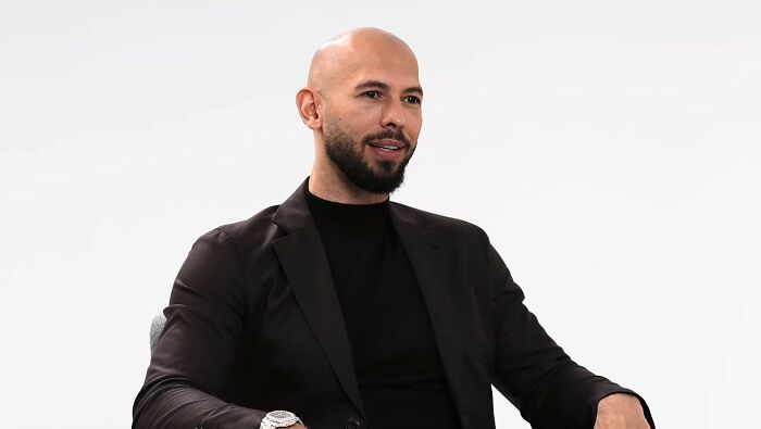 A bald man with a beard, wearing a black suit and turtleneck, sits against a white background. Manosphere content.