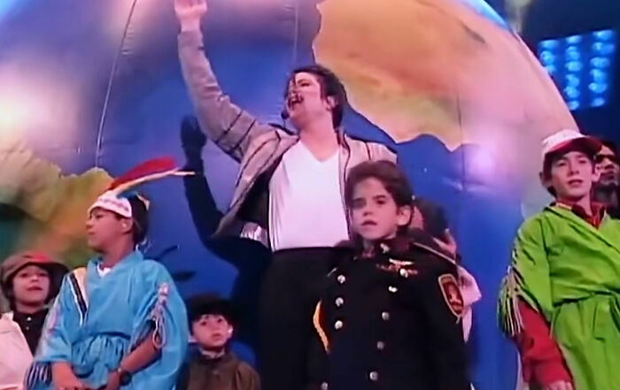 Michael Jackson on stage with children in various costumes, including one in a military uniform, against a large globe backdrop.