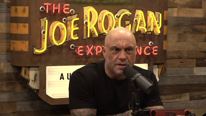 Joe Rogan speaking into a microphone. His show's neon sign, The Joe Rogan Experience, is visible in the background. Manosphere content.