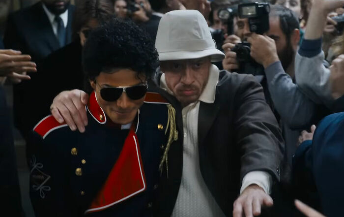 Michael Jackson impersonator in military jacket and sunglasses, escorted by a man in a hat through a crowd of paparazzi.