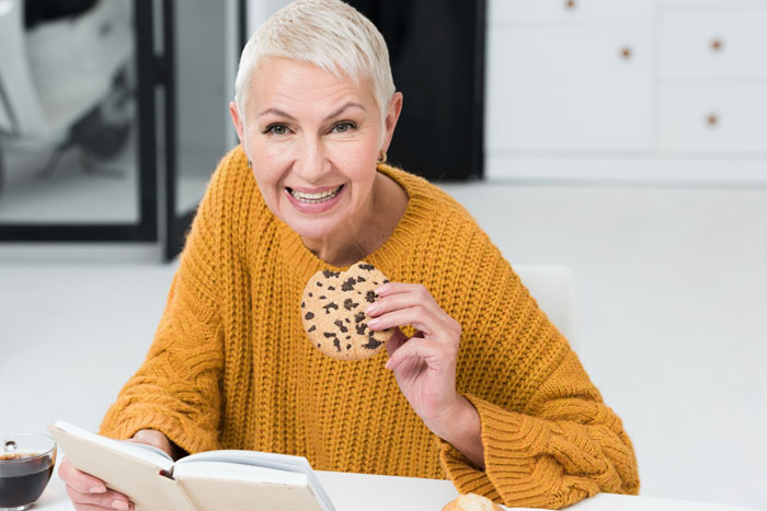 A smiling elderly woman in a yellow sweater holds a chocolate chip cookie, reading a book. She appears to be the MIL from the allergies story.