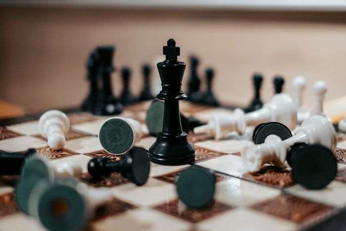 A black king chess piece stands victorious amongst fallen white and black pieces on a chessboard. Crows Hold A Grudge.