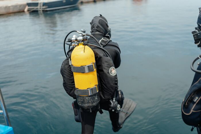 A diver in a black wetsuit with a bright yellow oxygen tank, about to enter the water. Disturbing d***h stories.
