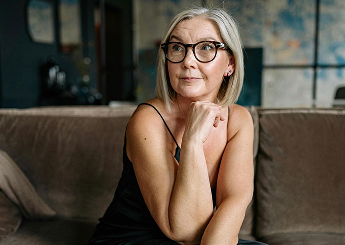 A thoughtful woman in glasses, with gray hair, sits on a couch, possibly reflecting on MILs creating chaos.