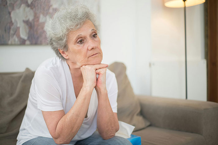 A thoughtful senior woman with gray hair sits on a couch, hands clasped. She is reflecting on MILs chaos.
