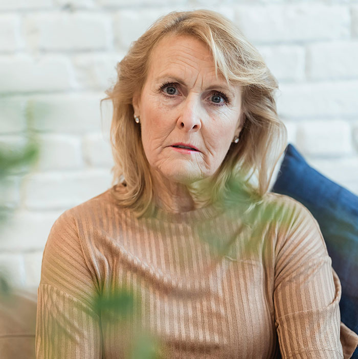 A serious-looking MIL, blonde with a beige top, creating chaos. The background shows a white brick wall and blue cushion.