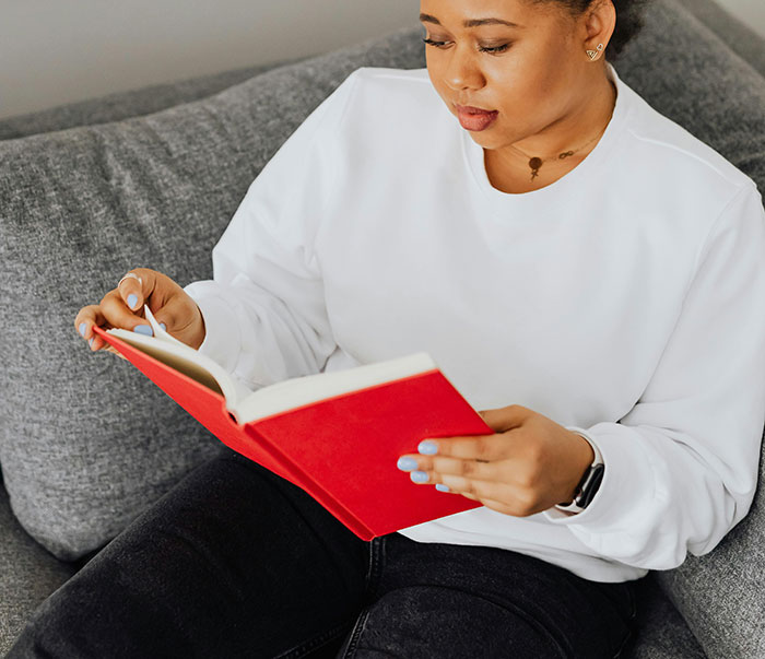 A woman in a white sweatshirt reads a red book. She could be reading about MILs and how they create chaos.