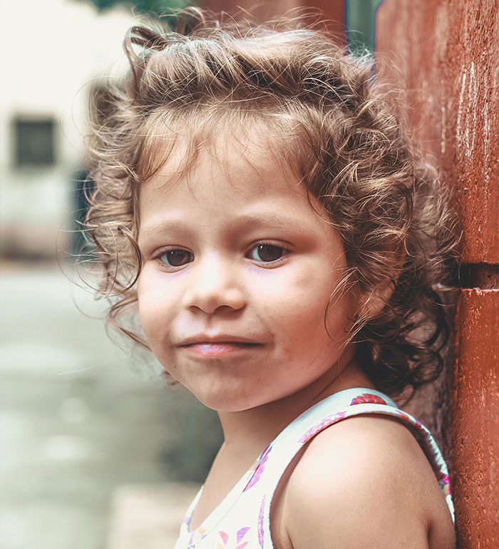 A cute little girl with curly hair, in a white tank top, looking at the camera, leaning against a red wall. MILs.