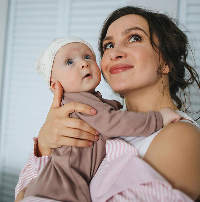 A smiling woman holding a baby, both looking up and to the left. The baby is wearing a light hat, innocent of MILs chaos.