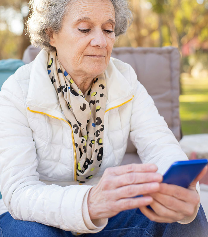 An older woman with grey hair, wearing a white jacket and patterned scarf, looking at her phone. MILs created chaos.