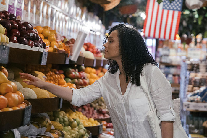 A woman touches fruit in a grocery store, an American flag in the background. MILs create chaos.