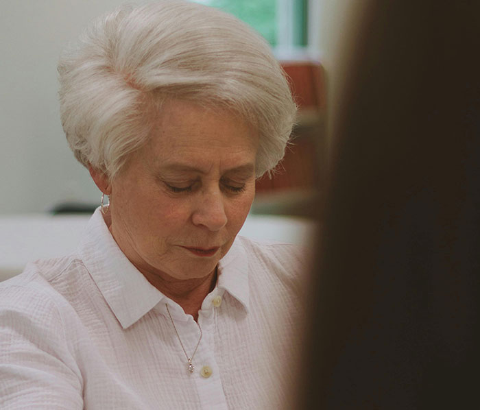 An older woman with white hair and a white shirt looks down, a common sight when MILs created chaos.