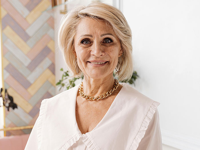 A smiling mature woman with blonde hair, wearing a white ruffled blouse and a chunky gold necklace, reflecting MILs creating chaos.