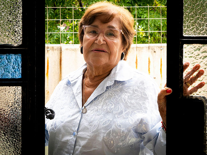 An elderly woman with brown hair and glasses looks seriously at the camera, framed by a doorway, creating MILs chaos.