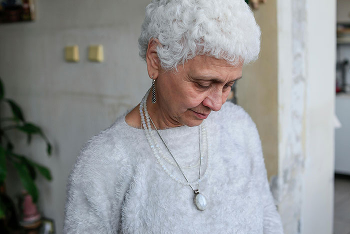 A contemplative elderly woman with curly white hair, wearing a fuzzy sweater and necklaces, possibly reflecting on MILs chaos.