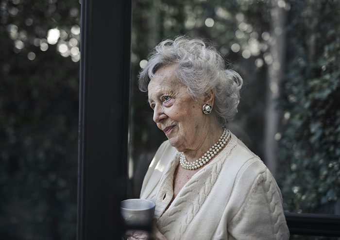 An elderly woman with gray hair, pearl earrings, and a triple pearl necklace, smiling gently while holding a cup. MILs created chaos.