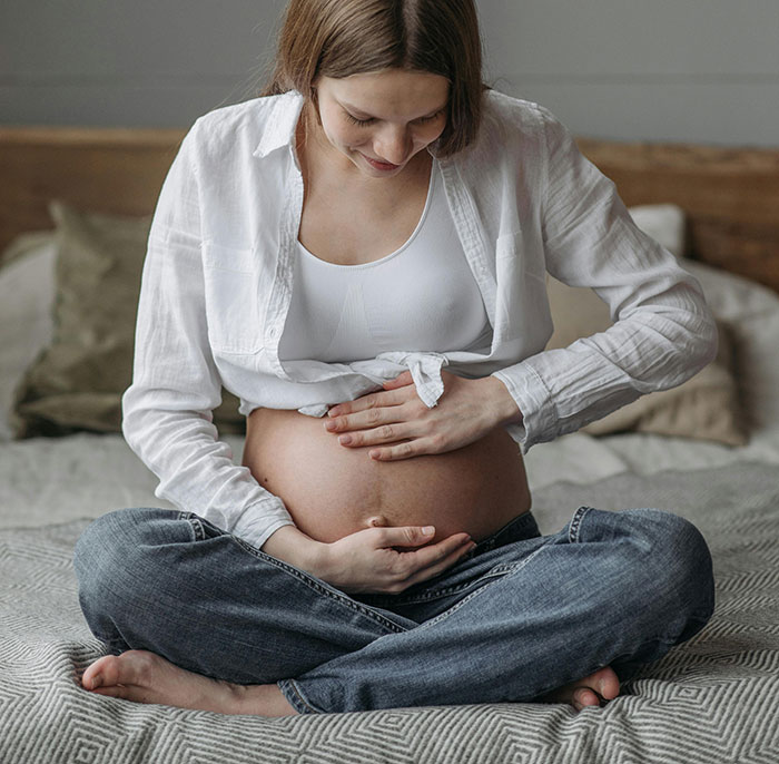 A pregnant woman in a white shirt and jeans, sitting cross-legged, gently touching her belly. MILs Chaos.