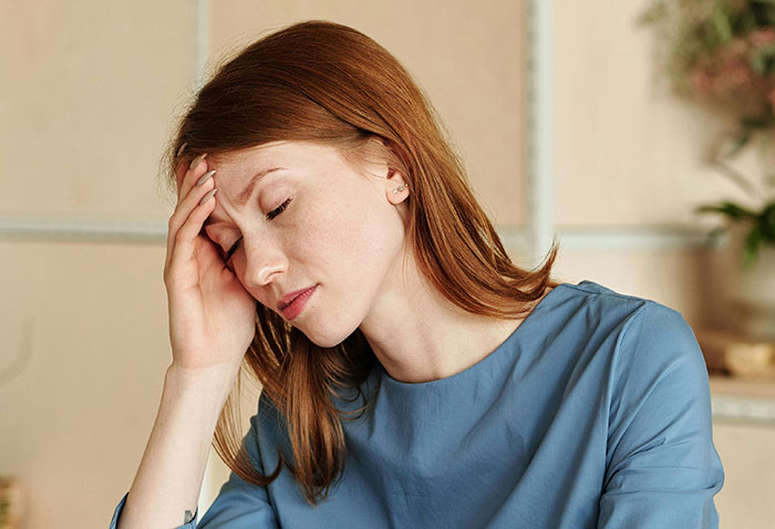 A woman with red hair and blue top holds her head, eyes closed, looking stressed, perhaps due to MILs creating chaos.