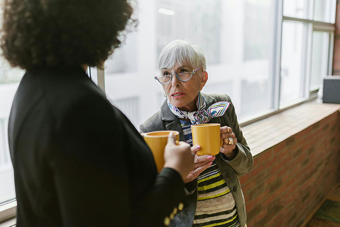 Two women with coffee mugs, one older with glasses and a scarf, talking about MILs creating chaos.