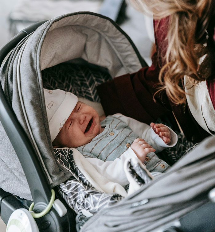 A crying baby in a car seat, with an adult leaning over, illustrating the chaos MILs can create.