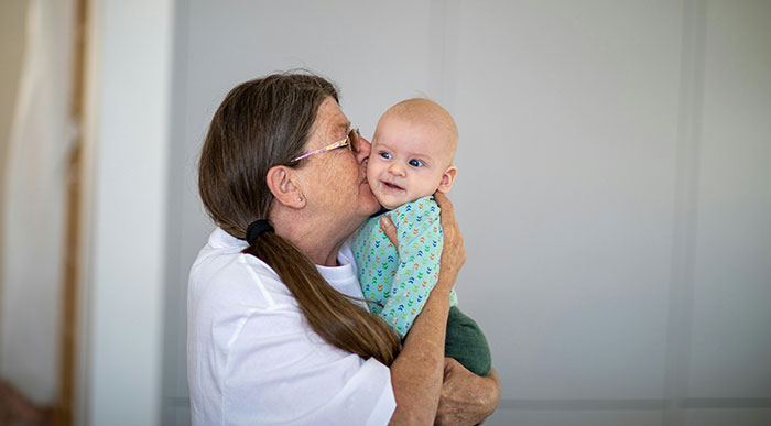 A MIL with long hair and glasses kisses a smiling baby on the cheek, creating joy where there could be chaos.