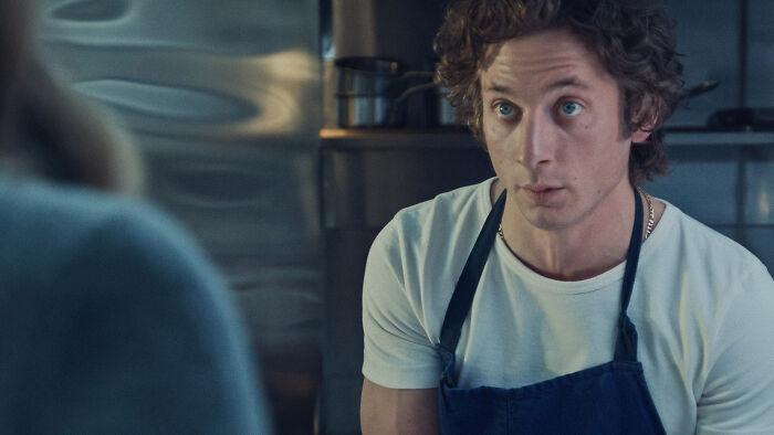 Chef Carmy da 5ª temporada de The Bear olhando atentamente. Ele veste uma camiseta branca e um avental azul na cozinha.