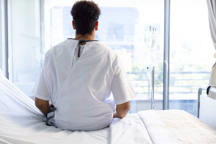 Patient in a hospital gown sits on a bed, looking out a window. Doctors are often amazed by how some are alive.