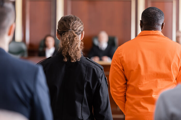 A prisoner in orange jumpsuit and a police officer standing in court facing a judge. Hardest cases for lawyers.