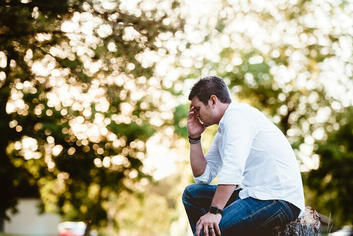 A man in a white shirt and jeans sits with his head in his hand, looking pensive. This image illustrates men suffering and manosphere content.